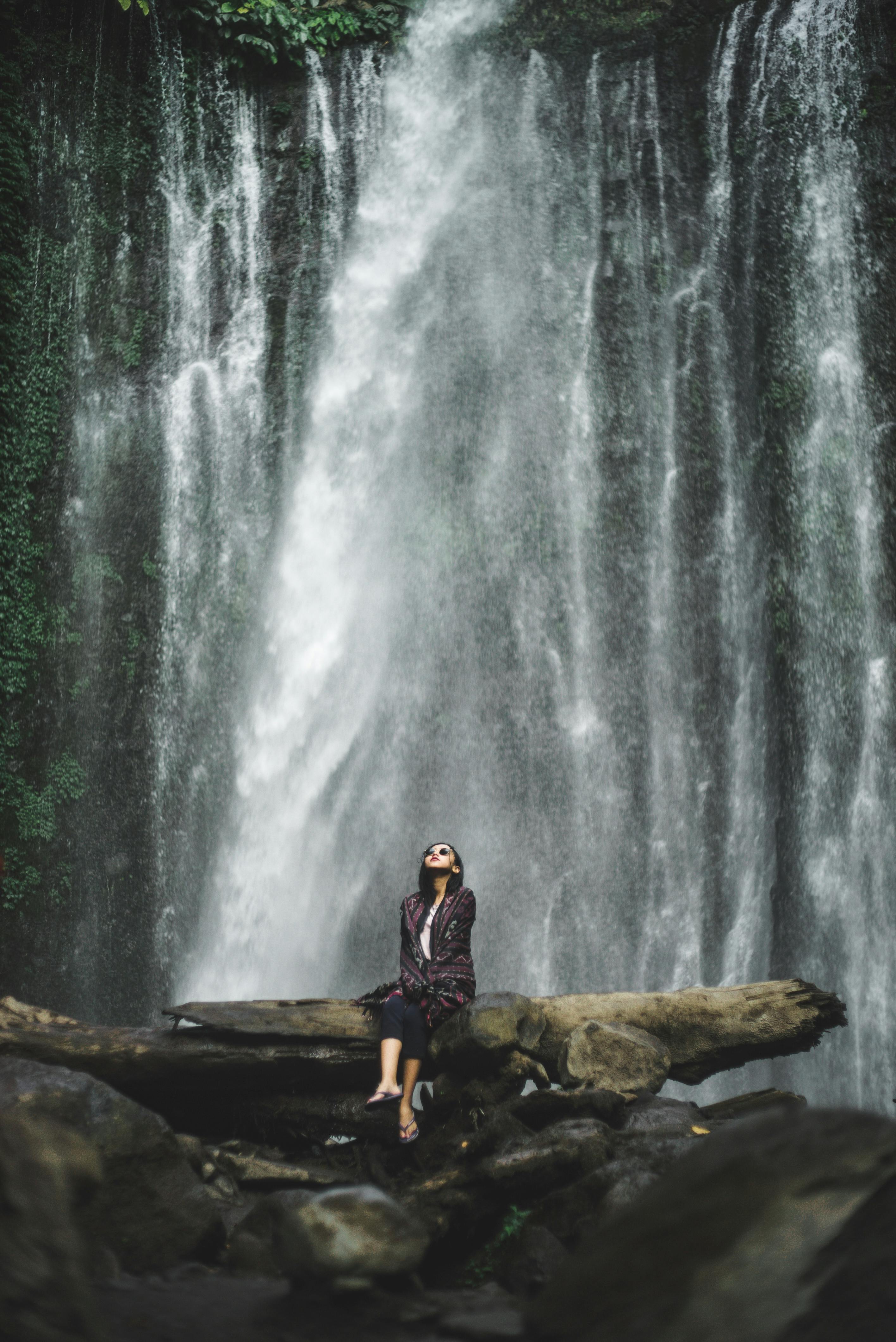 A woman seated below a vast waterfall, looking up, from doubt to awe in a single moment