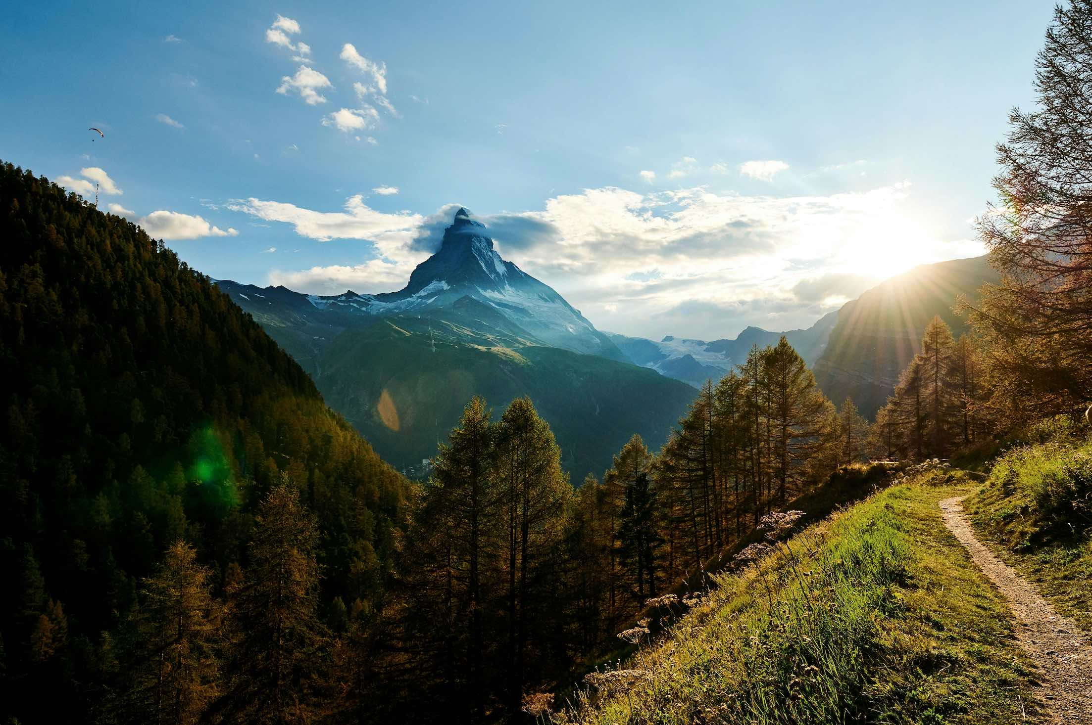 A picture of the sun peeking behind the Swiss Alps at the magic hour, a winding hiking path leading off into the mountains on the horizon.